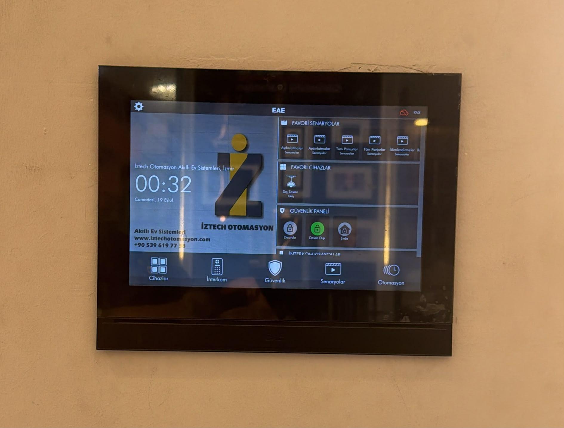 İzmir Jüpiter Elektrik showroom'unda duvara monte EAE Miola KNX dokunmatik panel; ekranda İztech Otomasyon logosu, favori senaryolar, güvenlik paneli ve interkom kısayolları